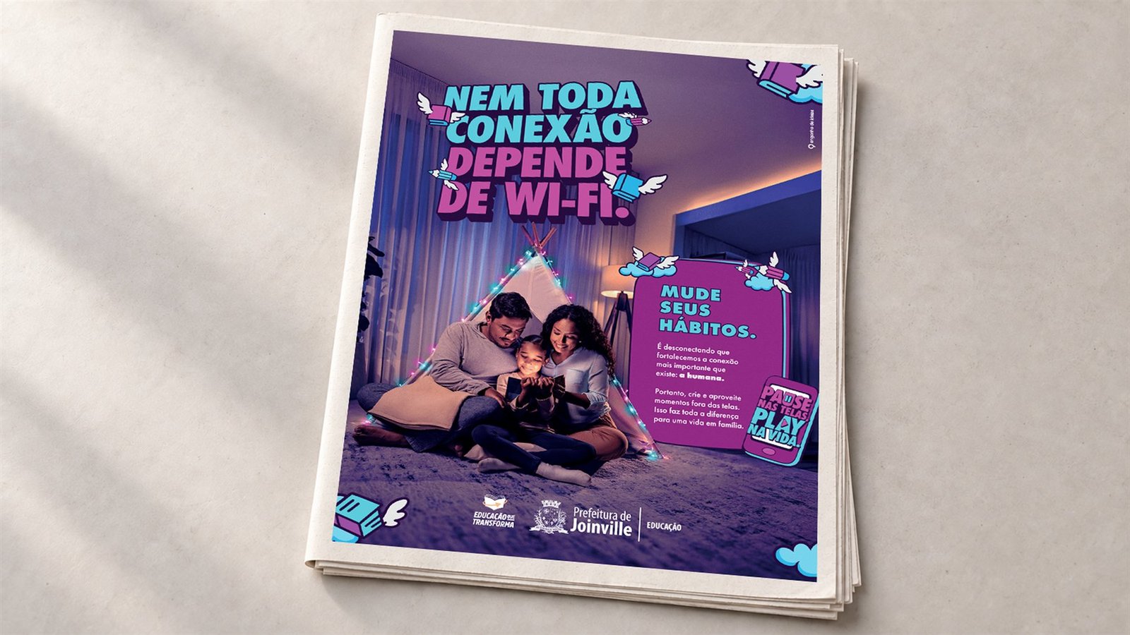 Anúncio de revista: pai, mãe e filha lendo juntos dentro de uma cabana iluminada por pisca-pisca, com o título “Nem toda conexão depende de wi-fi.” e o selo “Pause nas telas. Play na vida.”